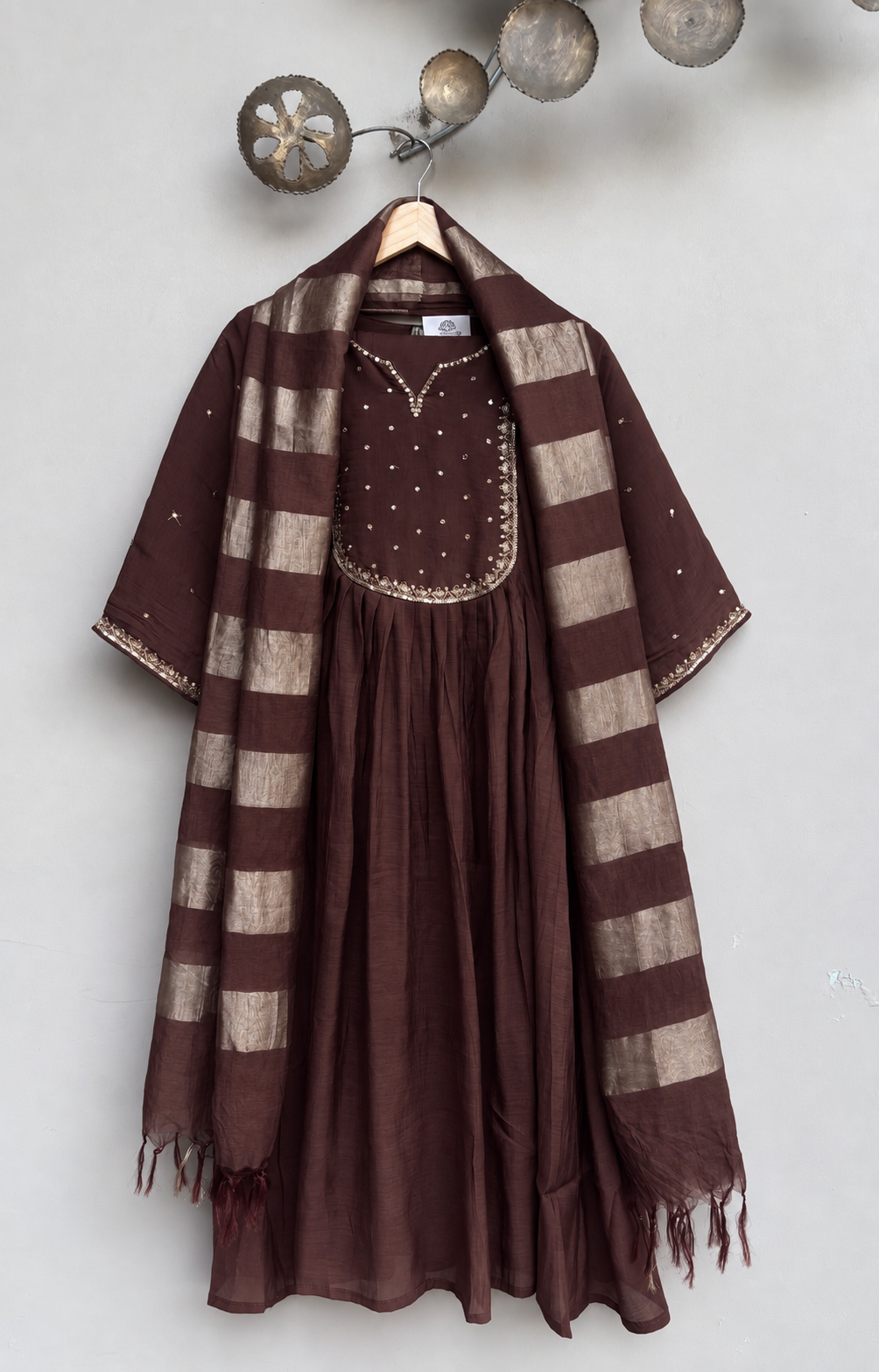 A brown and beige striped shawl draped over a brown embroidered kurta on a wooden hanger against a light gray wall.