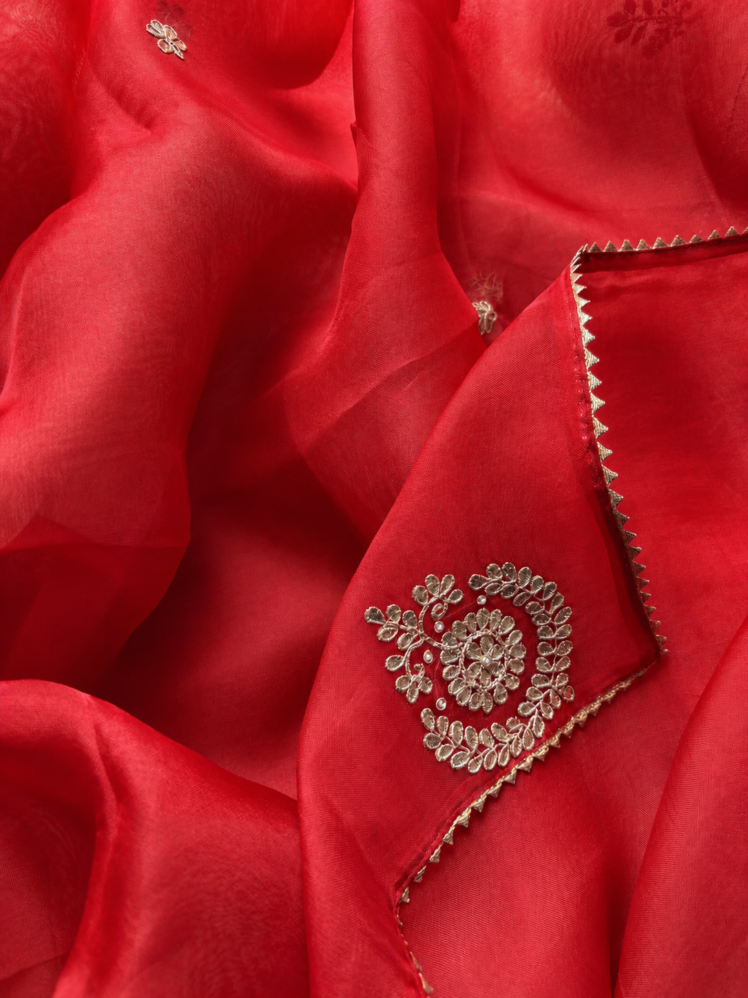 A close-up of a vibrant red fabric, likely silk or satin, featuring delicate gold floral embroidery and a zigzag gold border.
