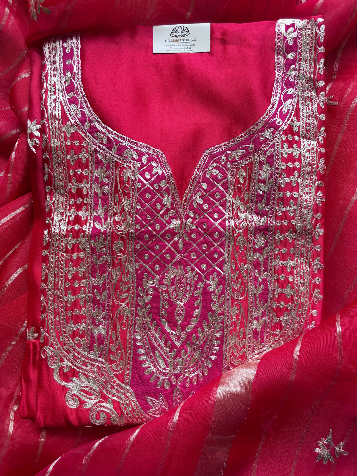 A close-up of a bright pink fabric garment, likely a kurta or blouse panel, featuring extensive silver mirror embroidery and a matching pink striped dupatta.