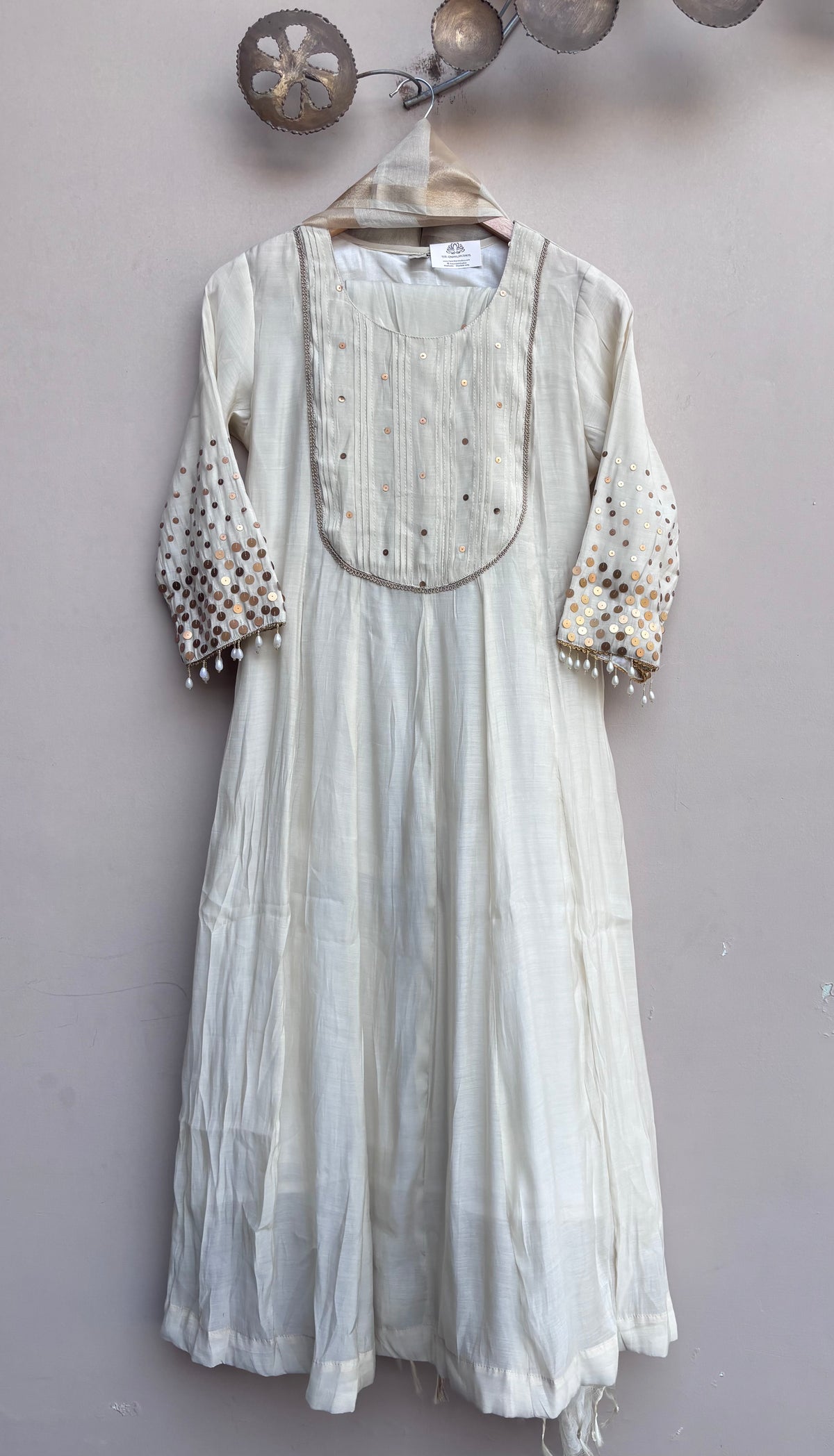 An off-white cotton dress with embroidered details on the chest and sleeves, hanging on a wooden hanger against a textured light gray wall.