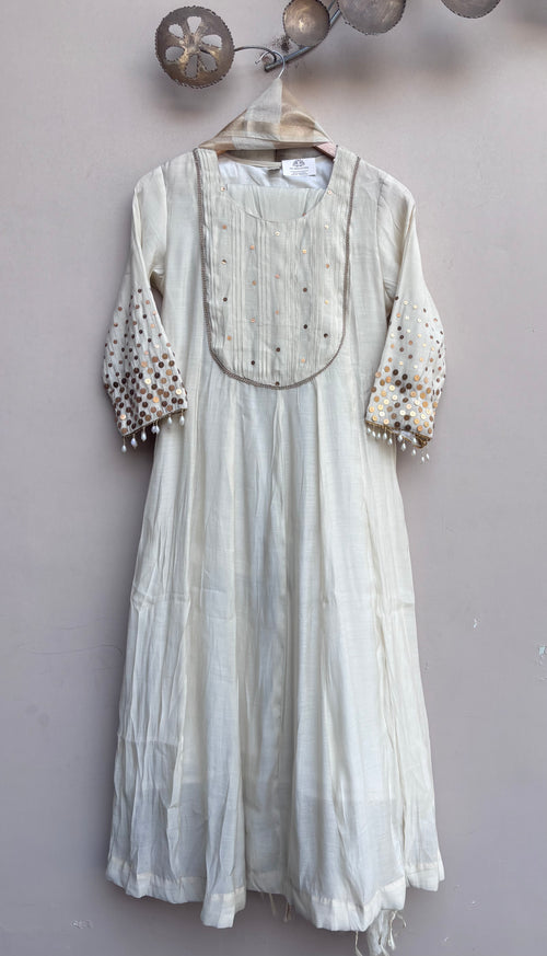 An off-white cotton dress with embroidered details on the chest and sleeves, hanging on a wooden hanger against a textured light gray wall.