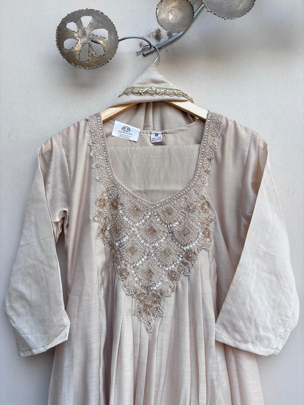 A light beige linen tunic dress with ornate gold and silver embroidery on the chest, hanging on a wooden hanger against a textured off-white wall.