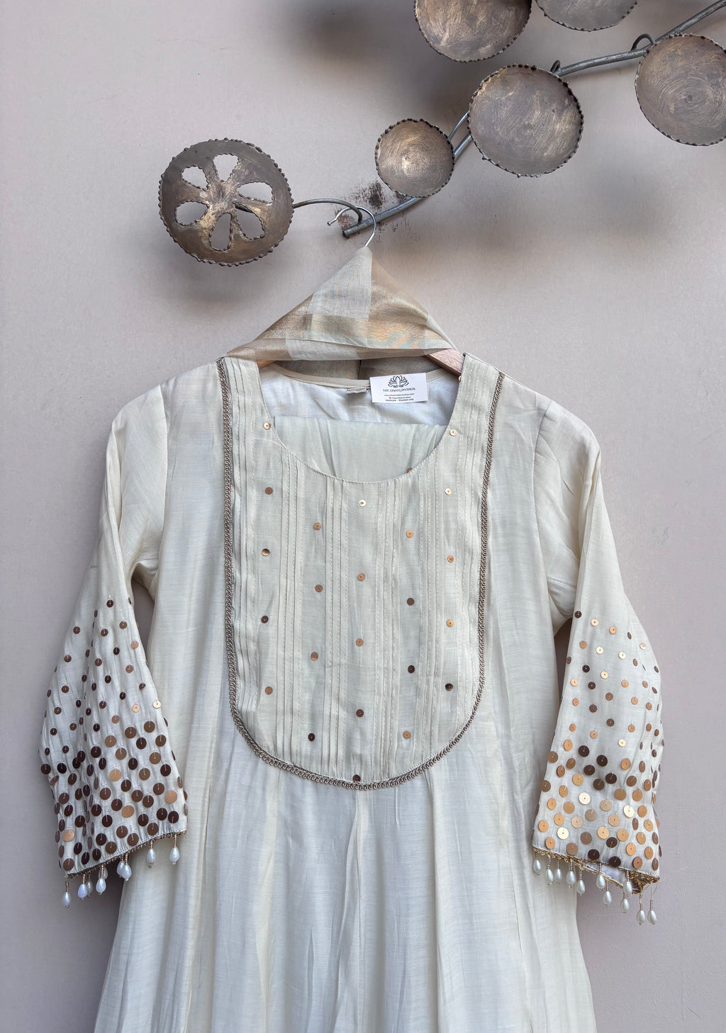 An off-white/cream long-sleeve tunic or kurta hanging on a wooden hanger against a light gray wall. The garment features a decorative embroidered yoke with small metallic dot details and matching embroidered cuffs with white tassel fringe.