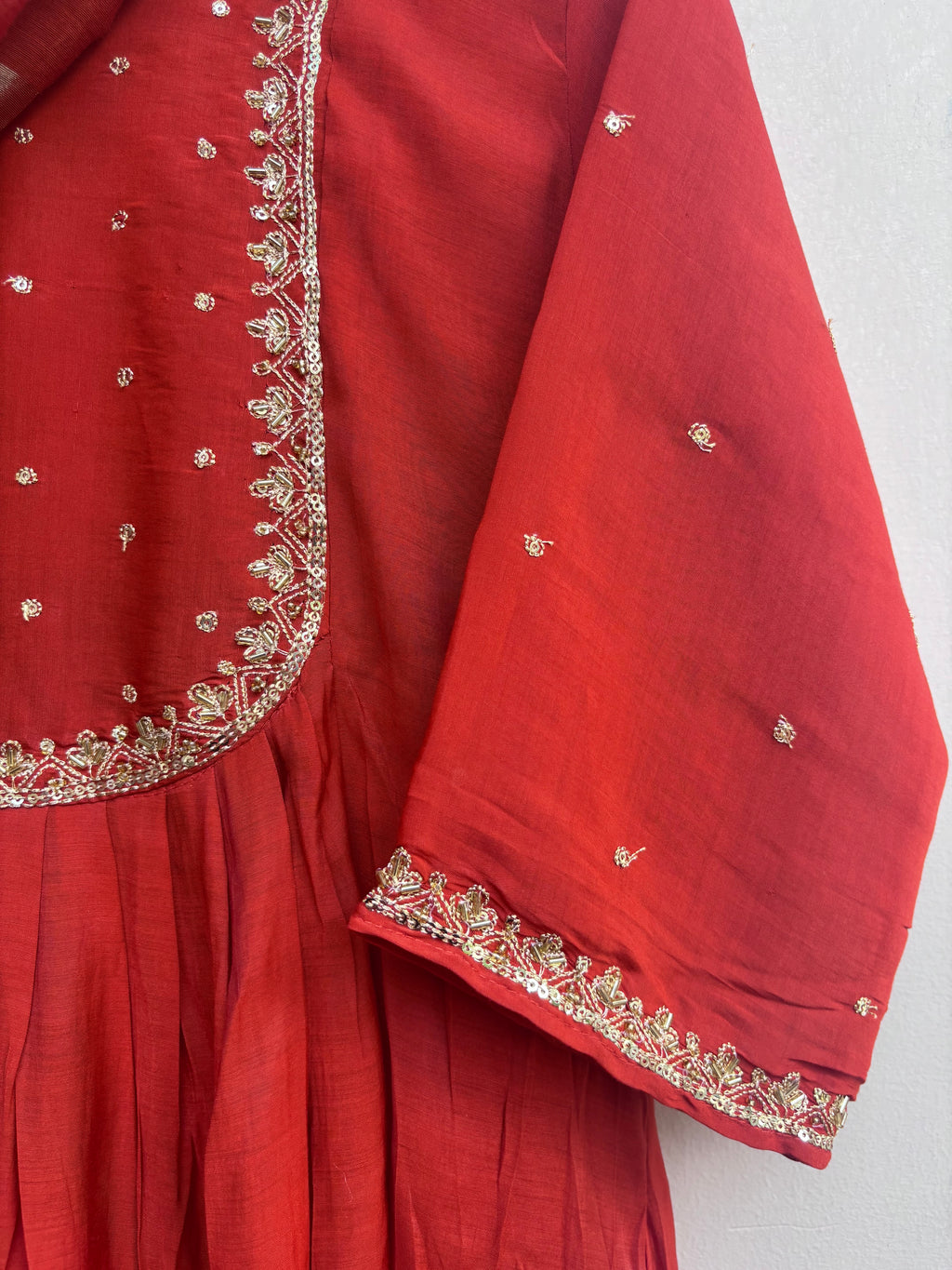 A close-up of a red traditional garment, likely a kurta or anarkali suit, featuring gold and silver embroidery along the border and scattered small metallic motifs on the fabric.