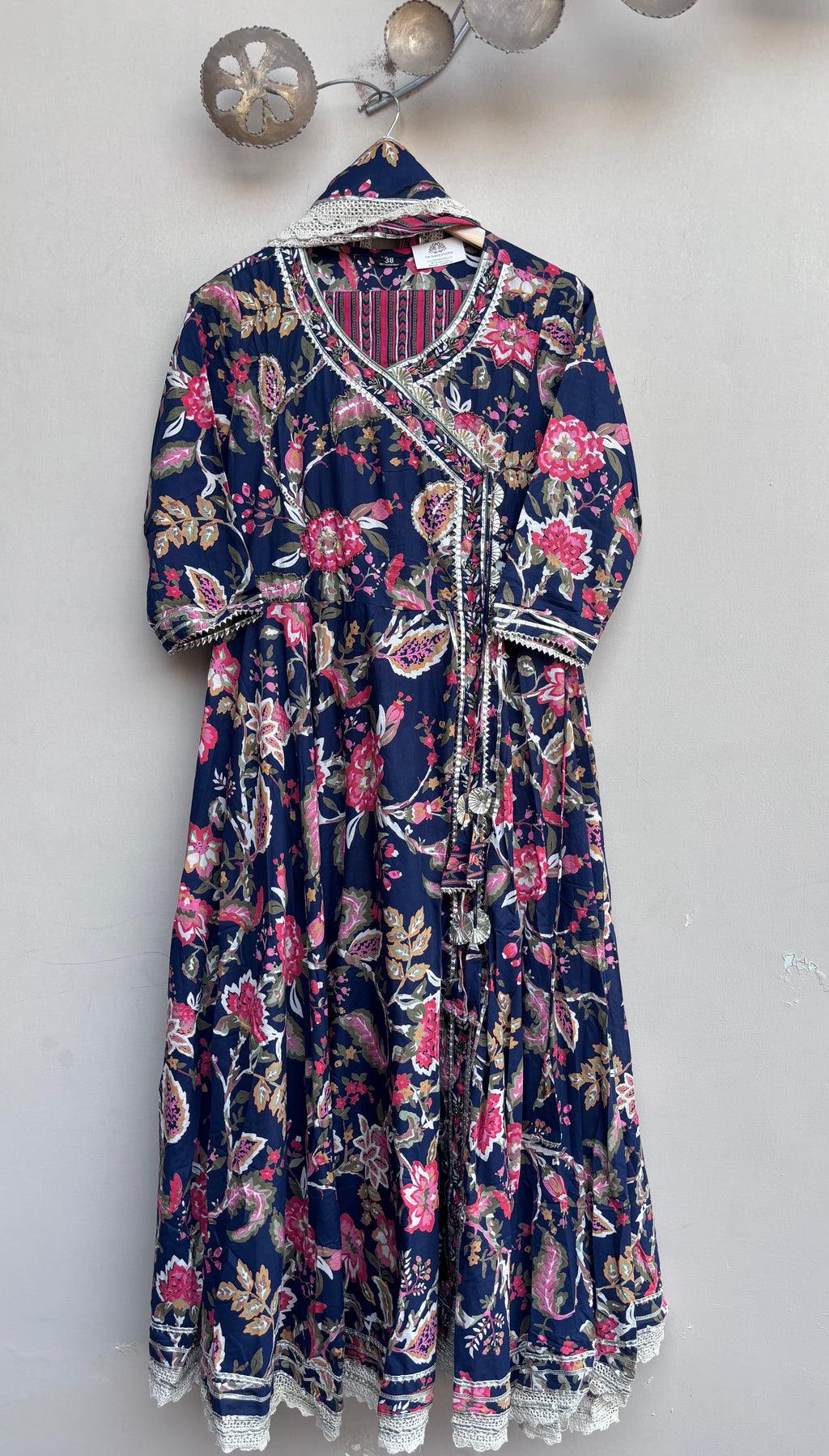 A navy blue maxi dress with a pink and red floral paisley print, featuring a V-neckline with white lace trim and a matching lace-trimmed waist tie, hanging on a wall.