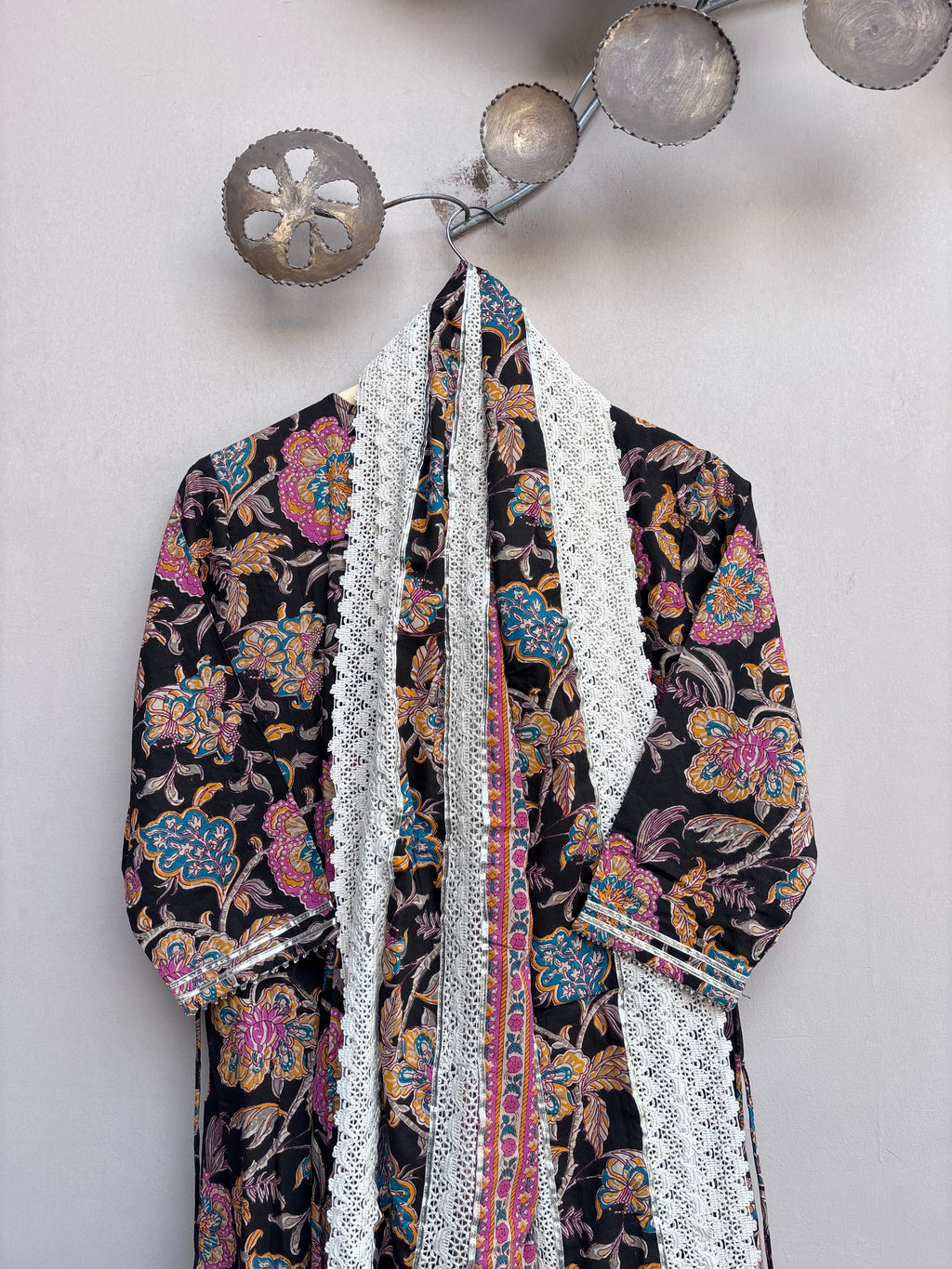 A long-sleeved printed jacket or robe with a black base and colorful floral motifs, featuring prominent white lace panels down the front and along the cuffs, hanging against a light gray wall.