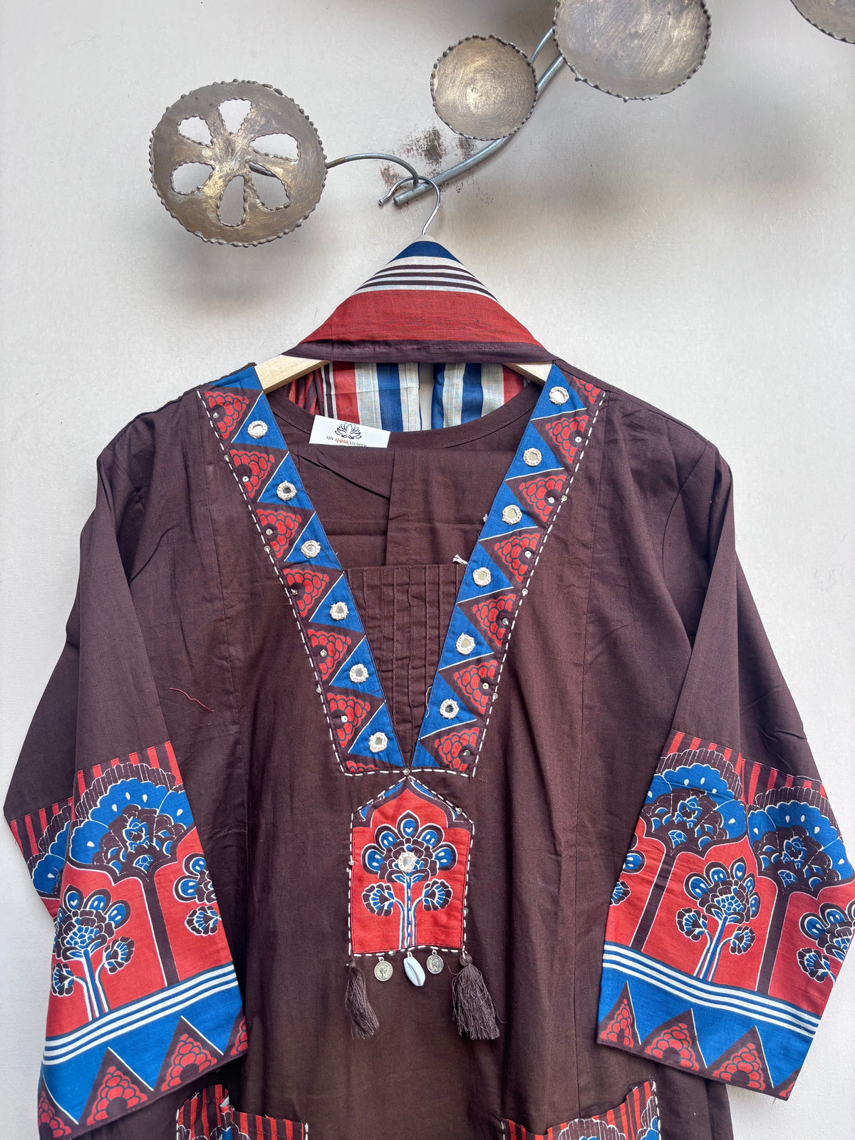 A brown cotton kaftan tunic with vibrant red and blue geometric embroidery, hanging on a wooden hanger against a light gray wall.