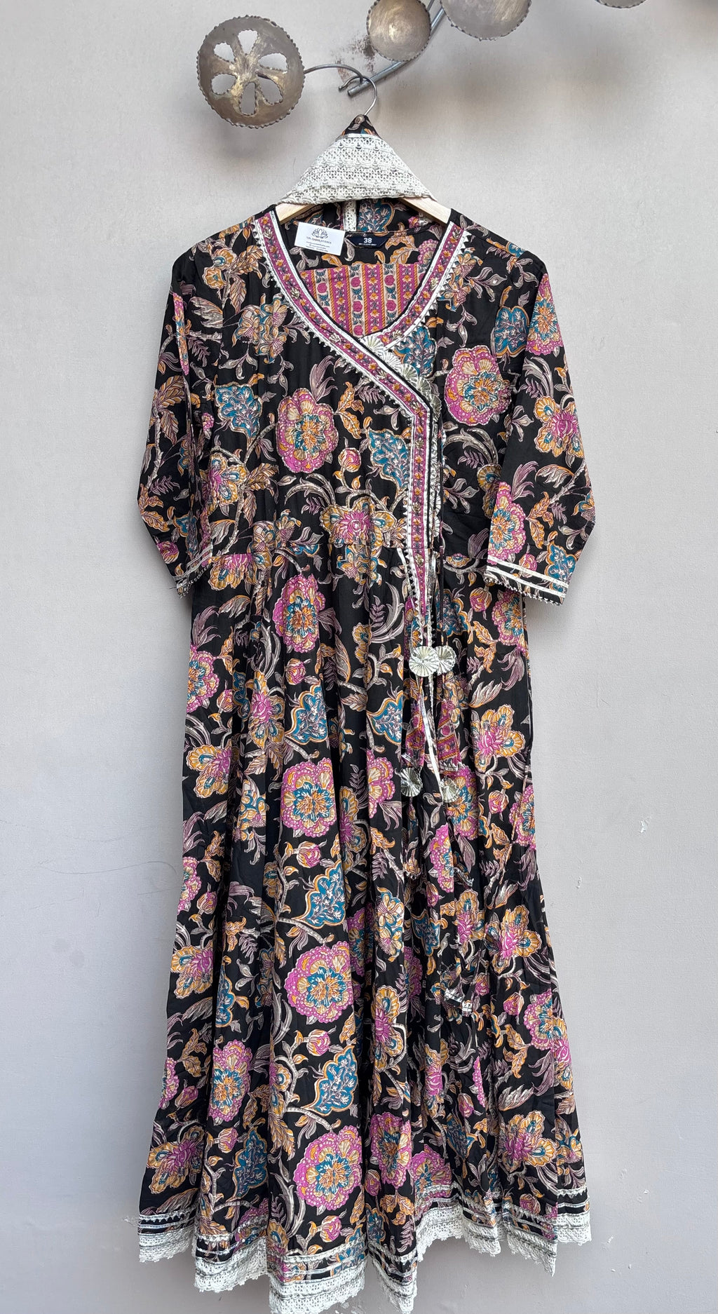 A black long-sleeve kaftan dress with a vibrant multicolor floral and paisley block print, featuring a white embroidered V-neckline and tassel tie, hanging on a white hanger against a light gray wall.