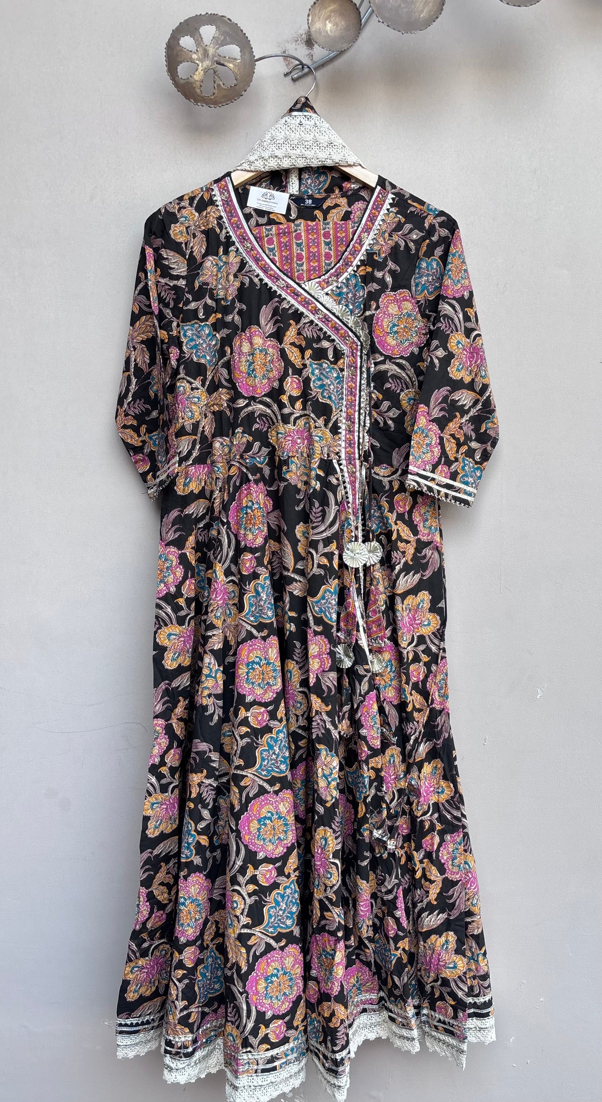 A black long-sleeve kaftan dress with a vibrant multicolor floral and paisley block print, featuring a white embroidered V-neckline and tassel tie, hanging on a white hanger against a light gray wall.