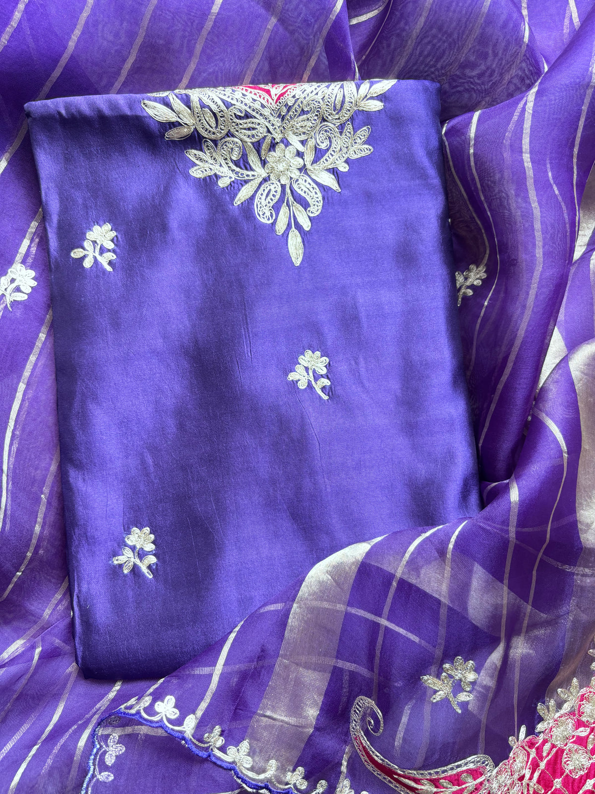 A folded purple fabric garment or textile with small white floral motifs and a large white embroidered floral design near the top, displayed on top of a matching sheer purple and white striped dupatta.
