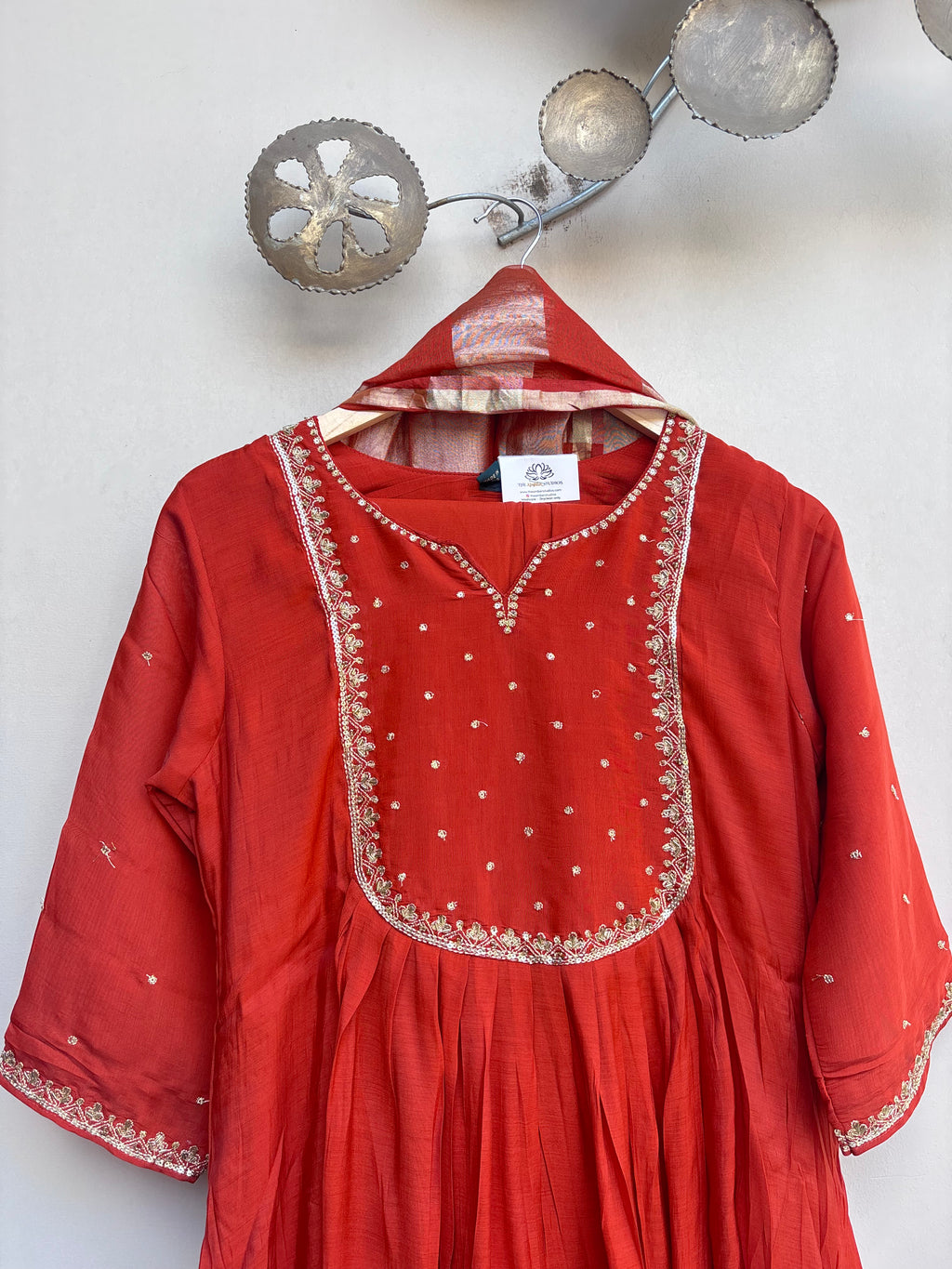 A red cotton kurta with gold embroidery and mirror work, hanging on a hanger against a white wall with decorative metal wall hangings.